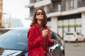 Fototapeta premium Confident young woman in a red coat stands next to a black car holding a smartphone and wearing sunglasses on a city street.