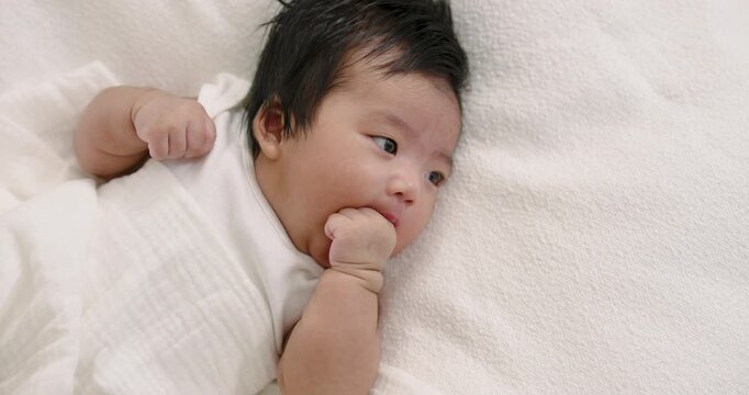 Asian newborn boy resting on white blanket gently sucking thumb while gazing forward with calm expression during quiet babyhood bonding moment at home