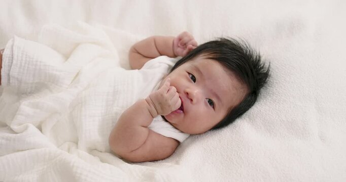 Asian infant lying on soft white fabric sucking tiny thumb with relaxed mood during quiet moment showing natural self soothing behavior of newborn baby in early stage of childhood development