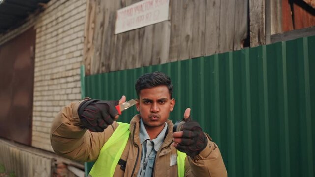 South asian construction worker in vest repairing fence with gloved hands, shows thumbsup and confident smile against green corrugated fence and brick shed, autumn light, practical jacket and tools,