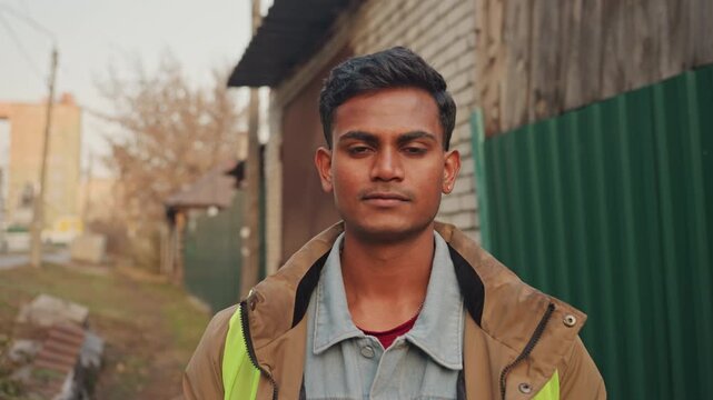 South asian man at fence, street fixer smiling during pause, community handyman near wooden shed, medium portrait with denim layers and reflective strip, approachable expression and sunset glow,