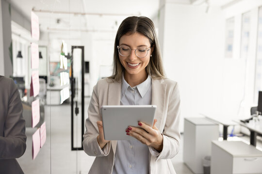 Young professional woman using digital tablet stands next to glass wall with sticky notes in contemporary office, accessing data, preparing presentation, making online task at workday. Tech, business