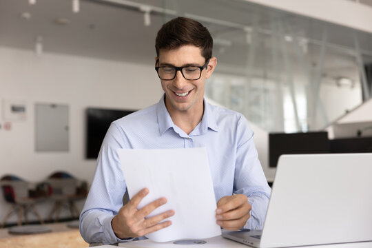 Businessman in casual shirt wearing glasses sitting at desk with open laptop in modern office, smiling, reads through printed document, reviewing good news, learns report with successful sales result
