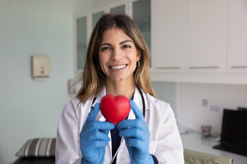 Obraz premium Head shot portrait of smiling Hispanic woman cardiologist dressed in white medical coat holding red heart object in her hands, posing for camera in clinic office. Heart health awareness, cardiology