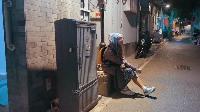 Nighttime urban solitude. Woman under streetlamp scrolling messages. Quiet city alley illuminated by neon light. Woman in casual attire sits alone in urban alley softly lit by neon glow