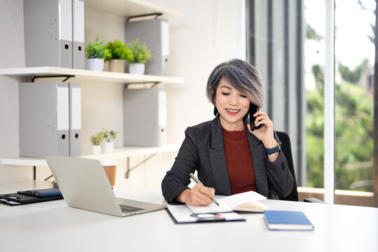 Happy smiling mature middle aged business woman, 50s professional lady executive manager talking on the phone making business call on cellphone and taking note at work in office .