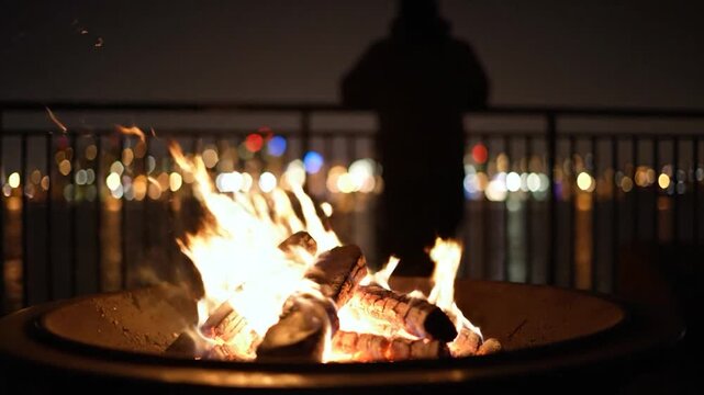 A close-up shot of a burning fire pit with flames and wood, with a blurred background of lights and a person
