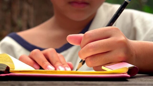 Close up of young Asian girl's hand writing in a notebook with a pencil, focused on studying in the park.