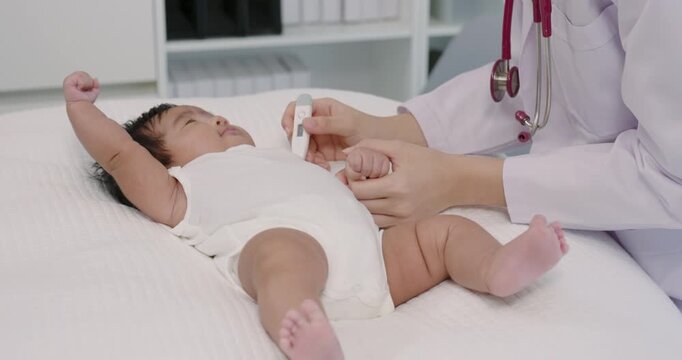 Asian female pediatrician gently places thermometer under newborn baby boy&rsquo;s arm during early clinical checkup showing soft caring routine in pediatric healthcare for infant temperature measurement