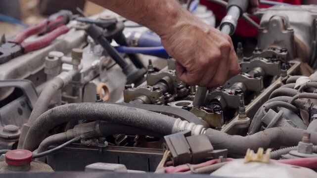 Mechanic works on engine components with focus and care. Hands adjust parts with precision, showing skilled attention. Engine bay is cluttered with wires and hoses