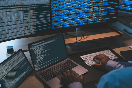 IT programmer working at a multi screen workstation with laptop and monitors displaying source code, showing software development workflow, coding process, technical productivity, and office computing