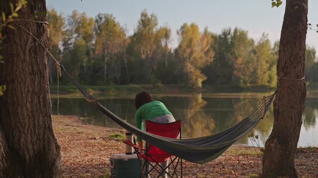 Woman white sitting in hammock lakeside packing cooler and chair then standing to fold hammock leaving empty hammock beside calm lake autumn trees reflecting in water solo camper enjoying peaceful