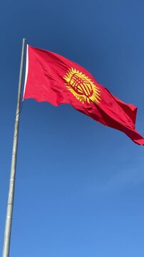 The national flag of Kyrgyzstan waves on a tall pole against a clear blue sky, its red field and golden sun symbol glowing brightly in the wind.