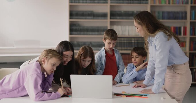 Group of diverse primary school pupils watching digital content on laptop in classroom during drawing class teamwork with teacher. Technology use, knowledge, creative educational activity, art project