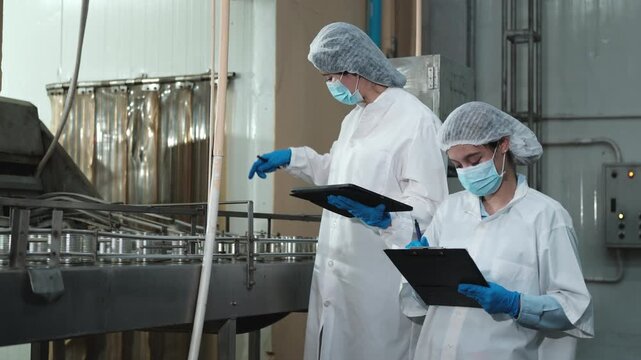 Two female industrial engineers inspect and examine quality control, production process at automate conveyor in canned food manufacturing factory, clean and protective uniforms for hygiene operations.