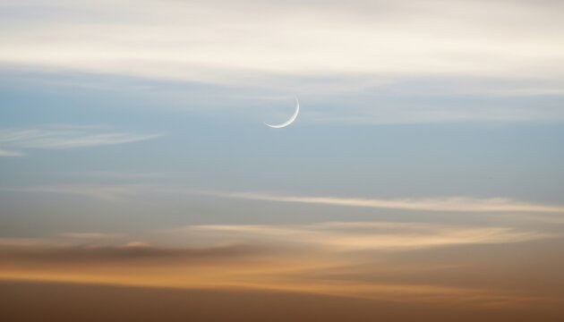 Atmospheric twilight sky with slender crescent moon appearing over a warm horizon for Ramadan Kareem greeting, Eid Al Adha religious banner and spiritual background