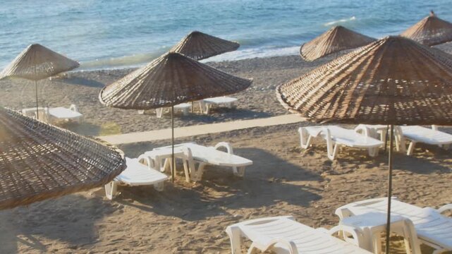 Empty beach resort with traditional wicker parasols and sunbeds overlooking the blue ocean waves. A serene morning scene capturing the quiet beauty of a coastal destination before the crowds arrive.

