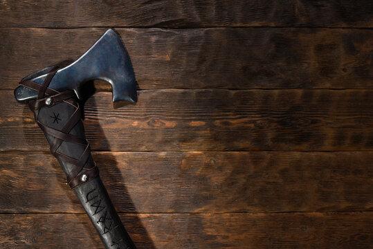 Forged steel axe with curved blade and leather wrapped handle engraved with runic symbols on wooden workbench table background. Top view.