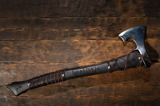 Forged steel axe with curved blade and leather wrapped handle engraved with runic symbols on wooden workbench table background. Top view.