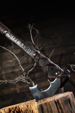 Forged steel axe with runic engraved handle wrapped in leather standing in wooden stump on dark rustic background. Front view.