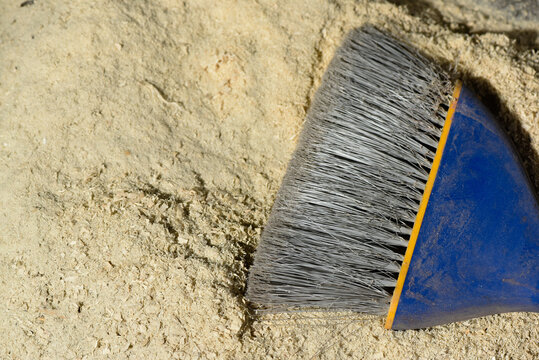 Hand broom with dense gray bristles cleaning fine sawdust on workshop surface background.