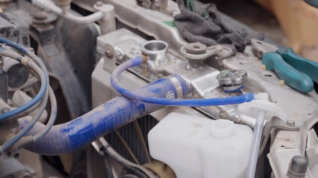 Water flows from a bottle into a coolant reservoir. Blue hose connects to engine system. Mechanic performs routine maintenance. Scene shows hands and tools actively working