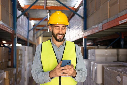 Smiling Hispanic male warehouse worker in uniform holding smartphone standing in logistics facility surrounded by storage racks. Supply chain management, digital inventory control, mobile apps use