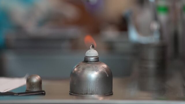 Alcohol burner in laboratory science chemistry equipment on metal table with dome cover and small flame, blurred background, experimental practical setup