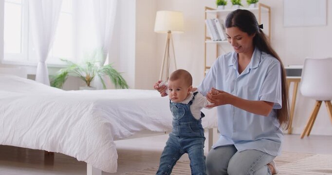 Loving mother helping small cute baby to stand, learn to walk holding hands, supportive and joyful moment, child achievement and care, development at home, balance, coordination act and move