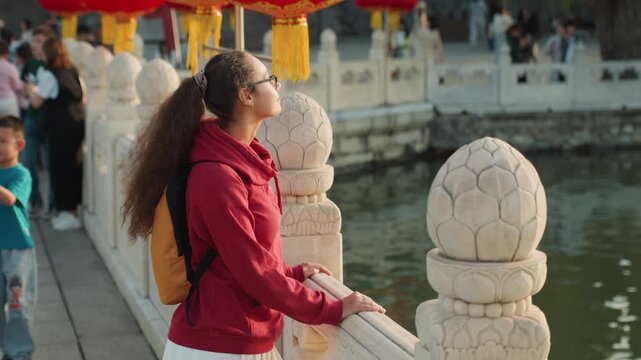 Young woman by waterfront bridge during lantern festival, wearing red jacket and yellow backpack, watching crowd and procession along stone railing, cultural costumes and tourists in background,
