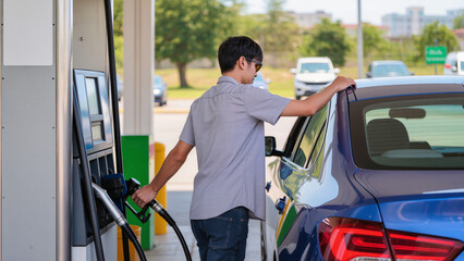 Man refuels blue sedan at station. ​Fuel pump nozzle held by driver. ​Person stands by blue car refueling. ​Gas station stop for car fuel. ​Refilling vehicle tank with gasoline nozzle. © Jakub