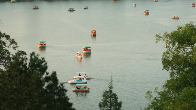 Calm lake dotted with colorful boats, families and friends on pedal boats and small motorboats, treelined foreground framing scene, distant buoys and soft hazy light, gentle ripples across glassy