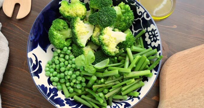 Fresh green vegetables like broccoli and asparagus in blue patterned bowl on wooden table. Healthy organic ingredients with peas prepared for steaming or salad in domestic kitchen. Concept of balanced