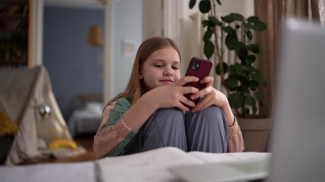 White girl scrolling smartphone on bed, knees hugged and laptop present, soft indoor light with tent and plant backdrop, casual smile while reading messages .