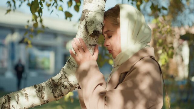 White woman praying by birch tree, eyes closed, hands resting on bark, urban park backdrop with blurred pedestrians, autumn light casting soft shadows, devotional mood, intimate closeup of quiet