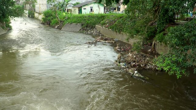 Handheld wide angle view of a highly polluted river with murky brown water and large amounts of floating plastic waste gathered near a residential riverbank in East Java, Indonesia.