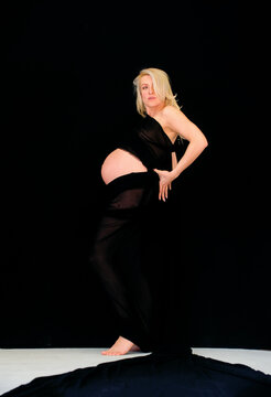 Pregnant woman in elegant black fabric posing against a dark background, highlighting the curved silhouette of her belly in a dramatic, artistic maternity portrait