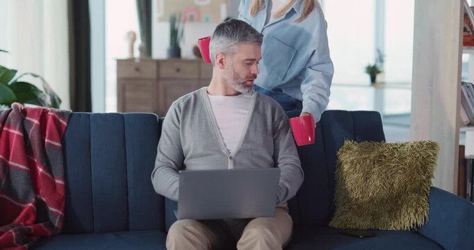 Caucasian attractive adult man working on laptop sharing coffee drinks with wife. Portrait of cheerful husband freelancing drinking coffeee smiling on camera.
