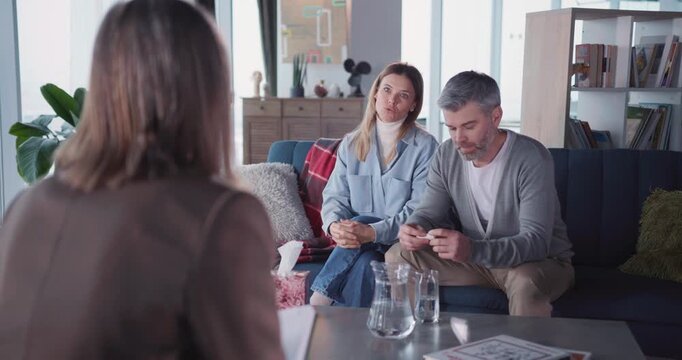Caucasian adult married family talking with mental health specialist female inside house. Wife complaining relationship issues sitting with husband on couch.