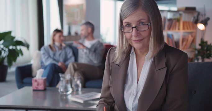 Portrait of Professional Mature Family Psychologist Female Wearing Glasses Writing Notes Posing to Camera while Consulting Adult Family Couple.