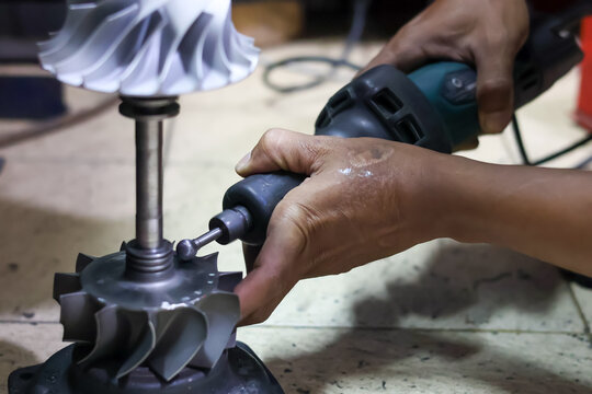 A close-up shot of a technician using a handheld electric rotary tool to grind and smooth a turbocharger turbine shaft in a professional workshop.