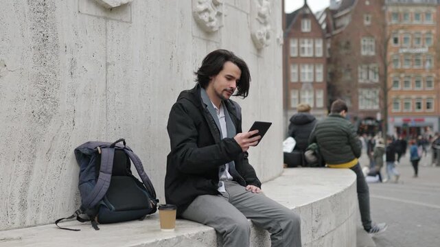 Young student reading an e-book while enjoying coffee outdoors in a bustling amsterdam square