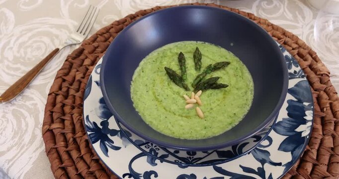 Top view of creamy green asparagus soup in blue bowl with floral pattern. Delicious healthy meal garnished with pine nuts and olive oil on wicker mat. Gourmet vegetarian dish served with fresh bread