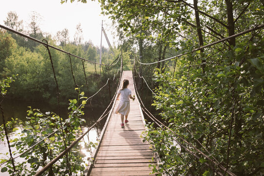 child girl walks on suspension bridge over river in nature summer outdoor. path from childhood