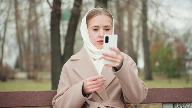 White woman on bench reacting to phone with dismissive gesture and stern expression, winter coat and scarf in leafless park, closeup of emotional confrontation and decisive reply to online provocation