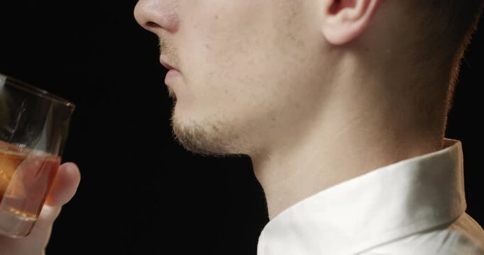 A close-up of a young bearded man in a white shirt sipping amber whiskey from a glass against a dark moody background.