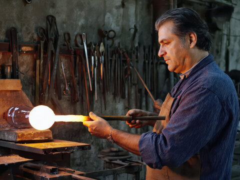 Glassblower in dark apron holding long pipe with glowing molten glass ball in warm workshop with orange furnace behind. Glass art craft concept