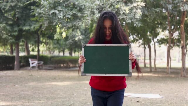 An Indian woman holding a blank green blackboard in front of her face while standing in a park. Creative concept image showing copy space for education, message display, or advertisement in a natural 