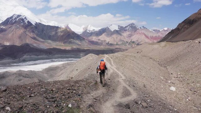 Lonely climber in mountaineering gear with backpack and trekking poles during Lenin Peak ascent, with mountain peaks in the background. Extreme active people, high-altitude mountaineering concept.