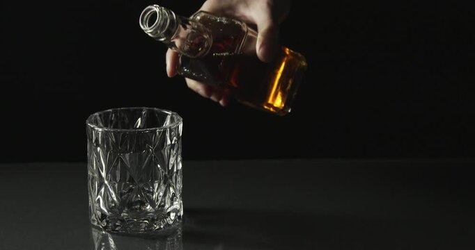 A hand pours whiskey from a bottle into a crystal glass in a dark cinematic scene.